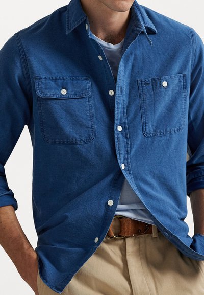 Blue button-up shirt made of soft fabric, featuring two chest pockets, white buttons, and rolled sleeves, paired with beige trousers.