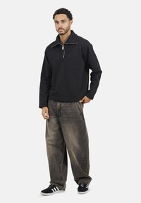 Pull noir avec un col montant et une fermeture éclair quart, associé à un jean gris délavé à coupe ample et des baskets noires avec des rayures blanches.