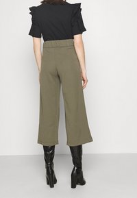 Kvinna som bär olivgröna vida cropade byxor, svart rufsig topp med korta ärmar och svarta knähöga klackstövlar, stående med ryggen mot kameran.