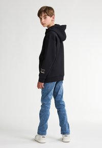 Sudadera con capucha negra de corte relajado, combinada con jeans de mezclilla azul claro y zapatillas blancas con logo. Diseño simple, con detalles mínimos.