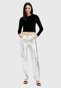 Fekete bársony, hosszú ujjú crop top fényes, fémes ezüst színű, széles lábú nadrággal párosítva. A modell keze a zsebében áll, fekete magassarkú cipőben.