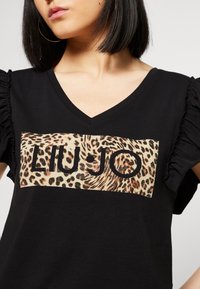 Zwarte V-hals T-shirt met ruches aan de mouwen, voorzien van een panterprint logo patch. De patch heeft een mix van bruine en beige tinten met pailletten.