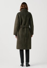 Olijfgroene, gestructureerde, lange jas met een brede kraag, een ceintuur in de taille en lange mouwen, gecombineerd met zwarte strakke broeken en hoge laarzen.