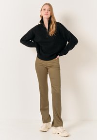 Schwarzer gestrickter Pullover mit einem Reißverschlusskragen, kombiniert mit taillierten olivgrünen Hosen. Cremefarbene Sneaker runden das Outfit ab.