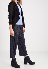 Czarny kardigan nałożony na jasnoniebieską bluzkę, zestawiony z teksturowanymi ciemnymi spodniami i czarnymi botkami na obcasie blokowym.