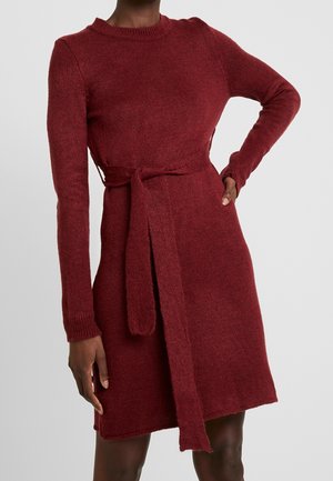 Abito di maglia bordeaux con maniche lunghe, scollatura rotonda e vita con cintura. La texture è morbida e leggermente pelosa, con polsini a coste.