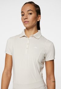 Ljusbeige polotröja med knäppning fram, krage och korta ärmar. Har en texturerad axeldesign och en liten vit logotyp.