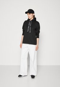 Felpa nera con logo bianco e stringhe, abbinata a pantaloni bianchi larghi e sneakers nere, completata da un cappellino nero. Tessuto morbido, vestibilità comoda.