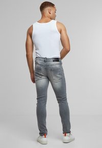 Férfi fehér ujj nélküli pólóban, szűk szürke farmernadrágban és fehér sportcipőben, narancssárga húzófülekkel, háttal állva szürke háttér előtt.