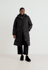 Zwarte pufferjas met een capuchon, voorzien van glanzende accenten en een volledige lengte. Draag het over donkere, wijde broek en zwarte schoenen.