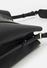 Borsa in pelle nera con una forma triangolare strutturata, caratterizzata da un accento in metallo dorato e una tracolla regolabile. Texture liscia, design minimalista.