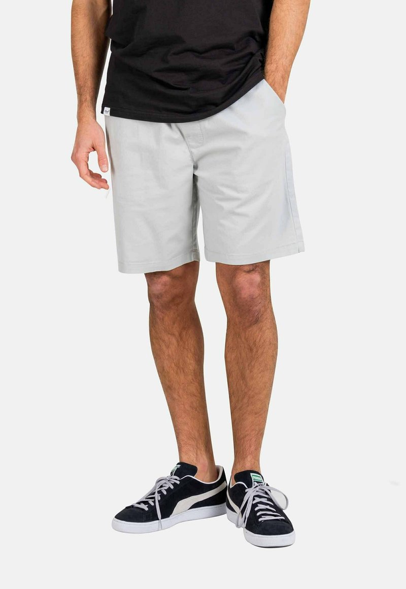 Pantalones cortos gris claro de tela, con un corte relajado y bolsillos laterales, combinados con zapatillas negras y una camiseta negra.
