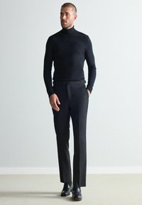 Schwarzer Rollkragenpullover mit figurbetonter Passform, kombiniert mit maßgeschneiderten schwarzen Hosen. Die Accessoires umfassen einen Gürtel und schwarze Halbschuhe.