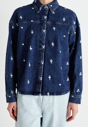Veste en jean bleu foncé avec petits motifs floraux blancs, devant boutonné, col et manches longues, portée avec un jean bleu clair.