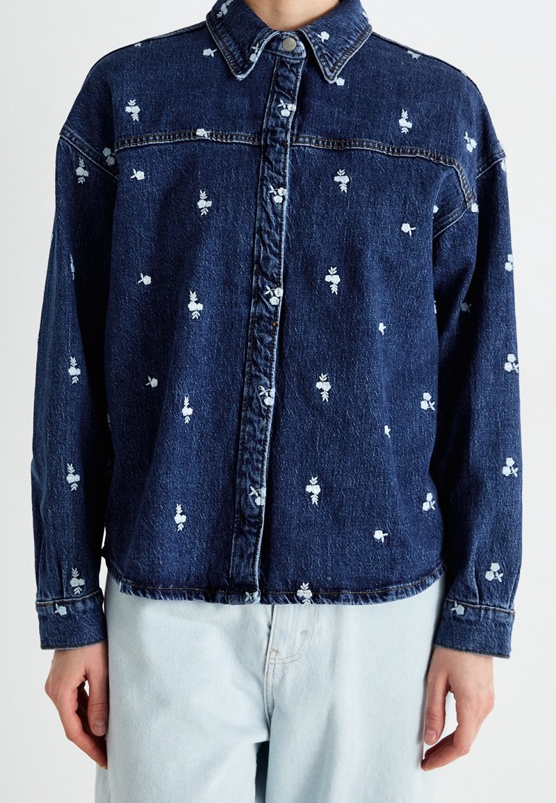 Veste en jean bleu foncé avec petits motifs floraux blancs, devant boutonné, col et manches longues, portée avec un jean bleu clair.