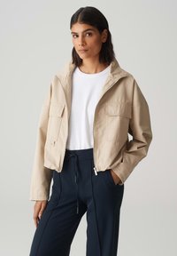 Beige cropped jack met een opstaande kraag, voorrits en twee borstzakken; gedragen over een wit shirt en donkerblauwe broek.