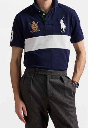 Homme portant un polo bleu marine avec une bande horizontale blanche, joueur de polo brodé et écusson, associé à un pantalon plissé gris foncé et une montre.