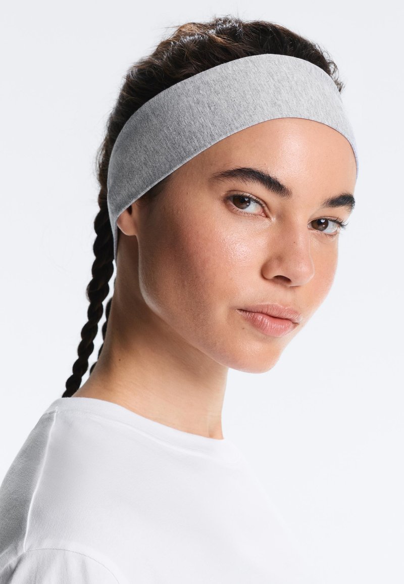 Femme aux cheveux tressés portant un large bandeau en tissu gris et une chemise blanche, regardant légèrement de profil sur un fond uni.