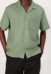 Camisa de botones de manga corta verde, hecha de un tejido ligero. Presenta un bolsillo en el pecho y un diseño de cuello relajado.