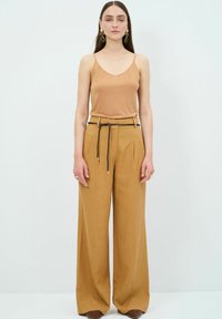 Beige satijnen camisole met dunne bandjes, gecombineerd met losse, tankleurige broeken met een gevlochten riem. Accessoires zijn ronde oorbellen.