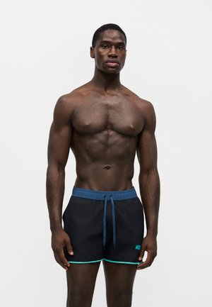 Hombre musculoso de piel oscura de pie, con pantalones cortos de baño negros con cintura azul y ribete turquesa, sobre un fondo claro y liso.