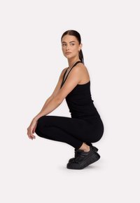 Kobieta w czarnym ubiorze sportowym i sneakersach przysiada, patrząc na bok, na tle jasnej, jednolitej ściany.