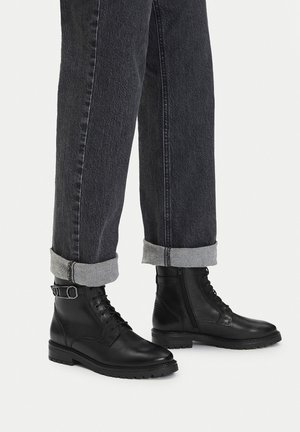 Czarne skórzane botki z okrągłym noskiem, sznurowane z przodu, z bocznym zamkiem i detalem w postaci klamry, zestawione z podwiniętymi ciemnymi dżinsami.