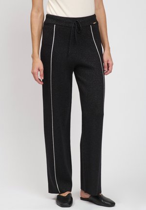 Person iført sorte bukser med vide ben med hvide sidelister, en hvid indstukket top og sorte slip-on sko, stående på en hvid baggrund.