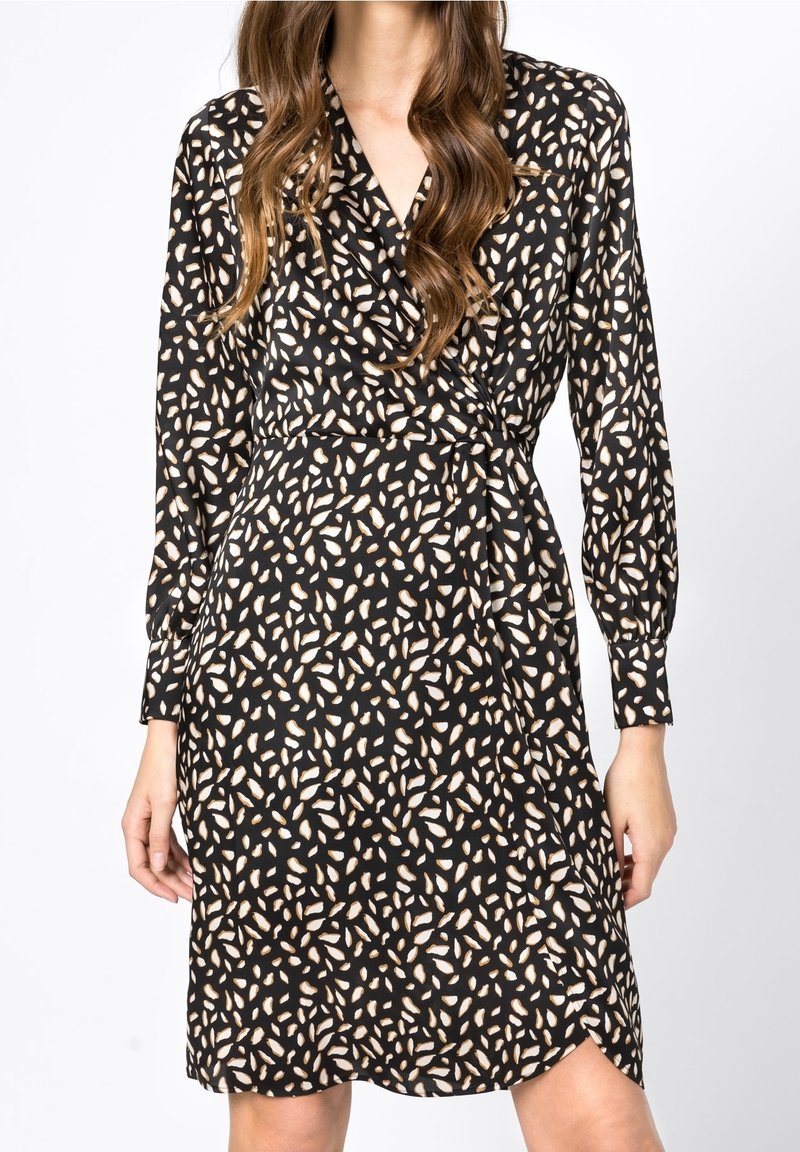 Femme portant une robe portefeuille noire avec un motif abstrait de feuilles beige, manches longues, et une légère fente devant.