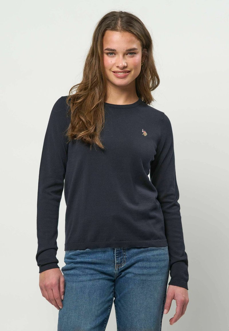 Tumši zila džemperis ar garām piedurknēm, klasisku apaļu kakla izgriezumu, ribotām aprocēm un mazu izšūtu detaļu uz krūtīm. Izgatavots no mīksta auduma.