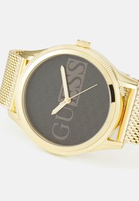 Goldene Mesh-Uhr mit rundem Gehäuse. Das dunkelgraue Zifferblatt zeigt das erhabene "GUESS"-Logo. Goldene Stunden- und Minutenzeiger, strukturierte Lünette.