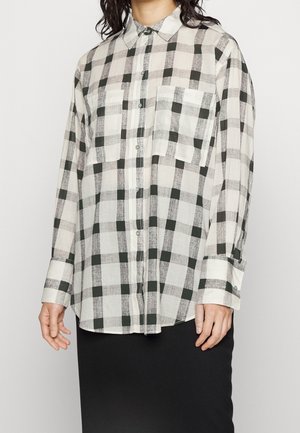 Persoon draagt een losse, lange mouwen blouse met een groot zwart-wit geruit patroon en twee borstzakken.