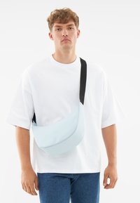 Borsa a spalla azzurro chiaro dalla forma curva, realizzata in materiale liscio, con una tracolla nera regolabile e un logo discreto sul davanti.
