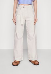 Calças largas de cor bege clara com uma cintura cingida, apresentando bolsos laterais e uma textura suave. Combinadas com sandálias castanhas com franjas.