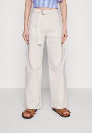 Pantalones de pierna ancha, de un beige claro y con cintura con cinturón, que cuentan con bolsillos laterales y una textura suave. Combinados con sandalias marrones de flecos.