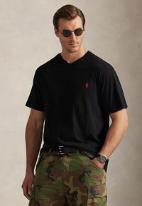 Man wearing black V-neck T-shirt with red logo, camo cargo pants, black belt, wristwatch, and round black sunglasses, posing with one hand in pocket.