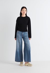 A young woman poses against a white background, wearing a black fitted top, wide-leg blue jeans, and gold clogs with decorative embellishments.