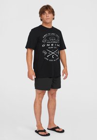 Sort bomulds t-shirt med hvid grafisk design, parret med mørke shorts og sorte flip-flops. Modellen står foran en helt hvid baggrund.