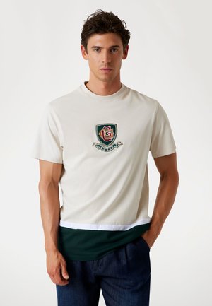 Cream short-sleeve T-shirt with a green and red embroidered logo at the chest, featuring a contrasting dark green lower panel and white trim.