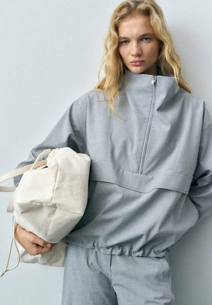Light grey zip-up jacket with high collar and elastic waistband, paired with a white drawstring bag. Smooth fabric with minimal design details.