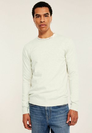 Mężczyzna o krótkich kręconych włosach, ubrany w kremowy sweter, niebieskie dżinsy, kolczyki koła i łańcuchowy naszyjnik, stoi na jednolitym tle.