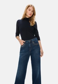 Weite Jeans aus dunkelblauem Denim mit hoher Taille, die mit dekorativen Nietenakzenten an den Vordertaschen und einer glatten Textur versehen sind.