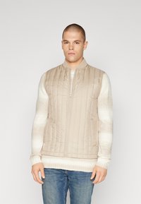Gilet matelassé beige avec col montant et fermeture éclair sur le devant, porté par-dessus un pull clair avec des manches rayées. Jean en denim décontracté.
