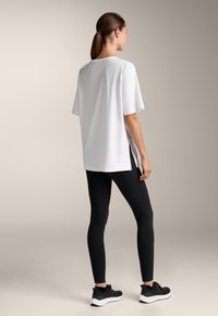 Hvid T-shirt med korte ærmer og sidespalter, parret med sorte leggings og sorte træningssko, sat mod en neutral baggrund.