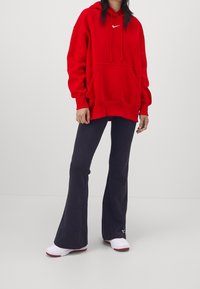 Sweat à capuche rouge avec une poche frontale et un logo blanc, associé à un pantalon évasé marine et des chaussures blanches avec des accents rouges. Tissu doux, design décontracté.