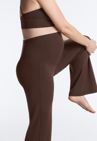 Personne enceinte étirant une jambe en portant des leggings de maternité marron et un soutien-gorge de sport assorti sur un fond blanc.