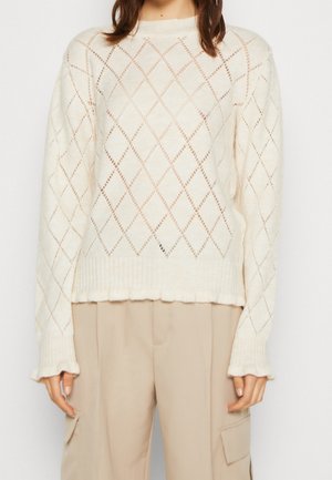 Woman wearing a white diamond-patterned open-knit sweater and beige high-waist pants with cargo pockets, standing against a plain background.