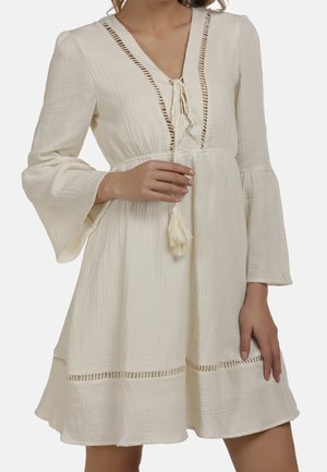 Femme portant une robe texturée couleur crème avec manches cloche, encolure à lacets, lien à pompons et détails de coupe ajourée.