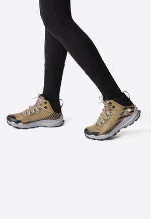 Person iført beige og sorte vandresko med sorte sokker og sorte leggings mod en ensfarvet lys baggrund.