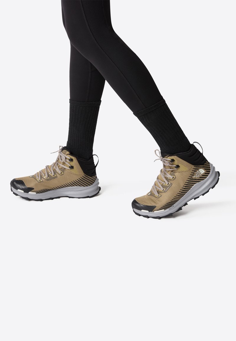 Personne portant des chaussures de randonnée beige et noir avec des chaussettes noires et des leggings noirs sur un fond clair uni.